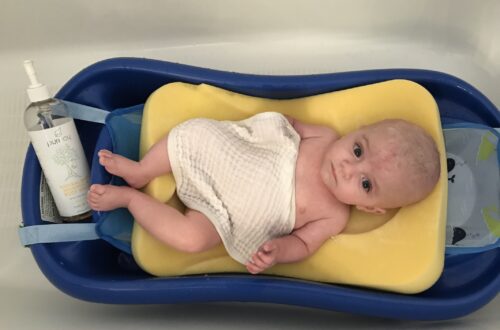 first years sure comfort deluxe baby tub with baby inside of it with a sponge sling and shampo caddy